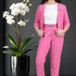 Young girl wearing a vibrant pink corduroy 3-piece suit including a blazer, trousers, and a silver-buckle belt, standing next to a white orchid plant.